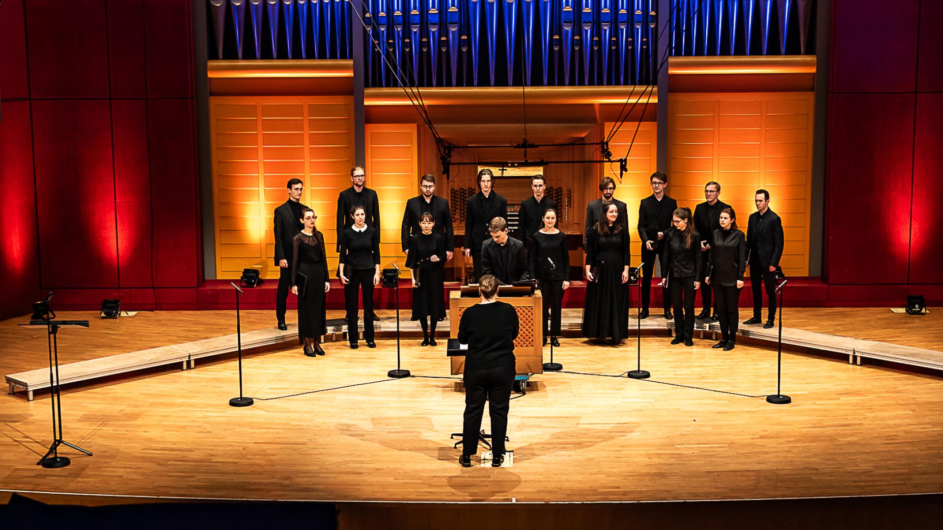 Das sechzehnköpfige Vokalensemble vor dem Orgelprospekt der Konzertsaalorgel. Mit dem Rücken zum Publikum steht die Dirigentin. Umgeben vom Vokalensembles ist der Studierende an der Truhenorgel.