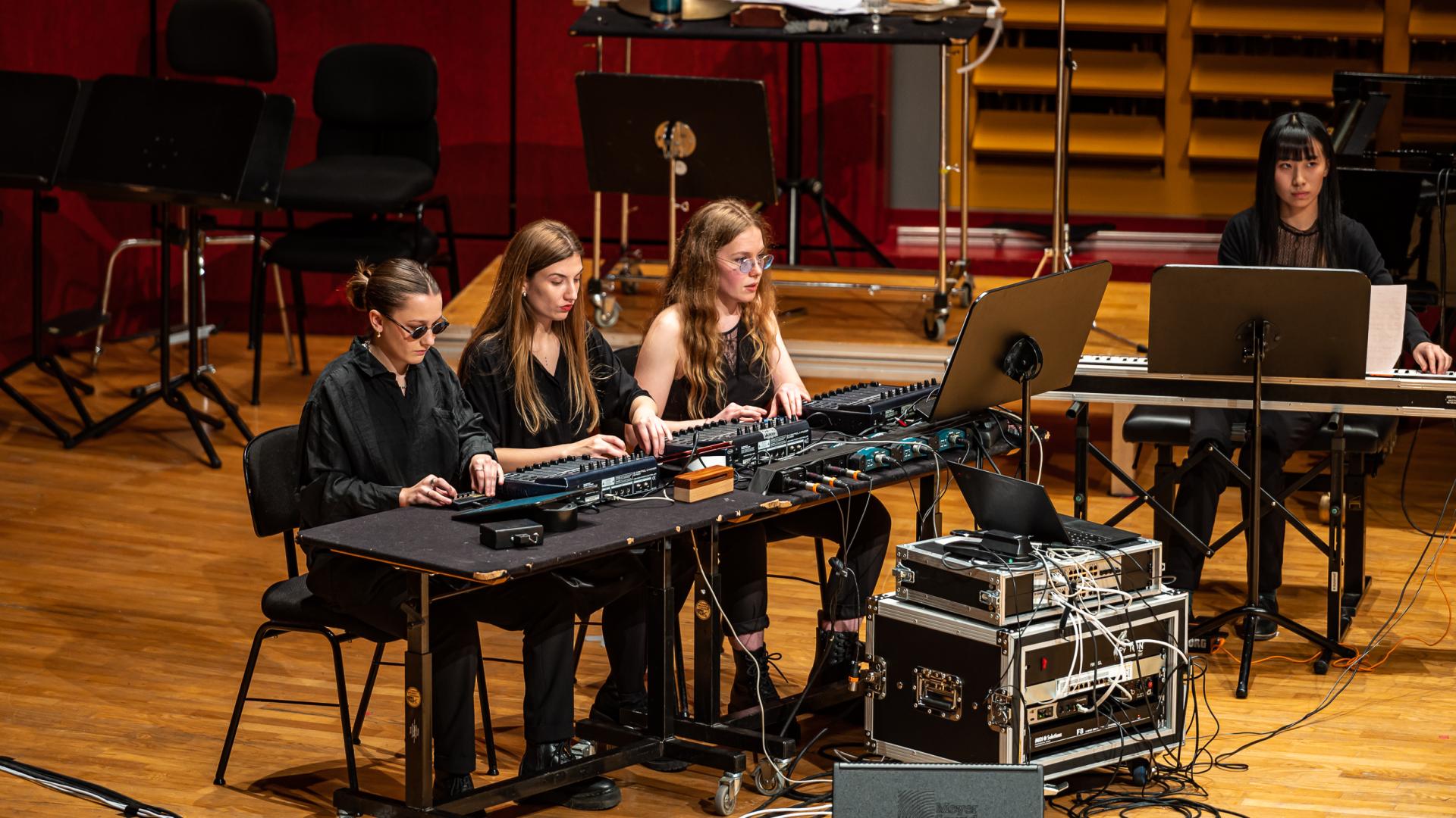 Dret Studierenden sitzen an einem Tisch mit unterschiedlichen Pulten. An einem gesondert Tisch sitzt eine Studierende mit Keyboard. Zwischen den Tischen sind Dutzende von Kabeln verlegt.