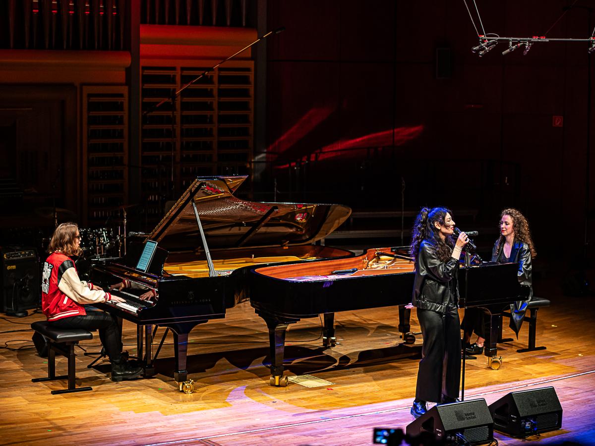 Schulmusikkonzert 2024: zwei Studentinnen am Flügel, die sich einander gegenüber stehen, eine Studentin, die singt.