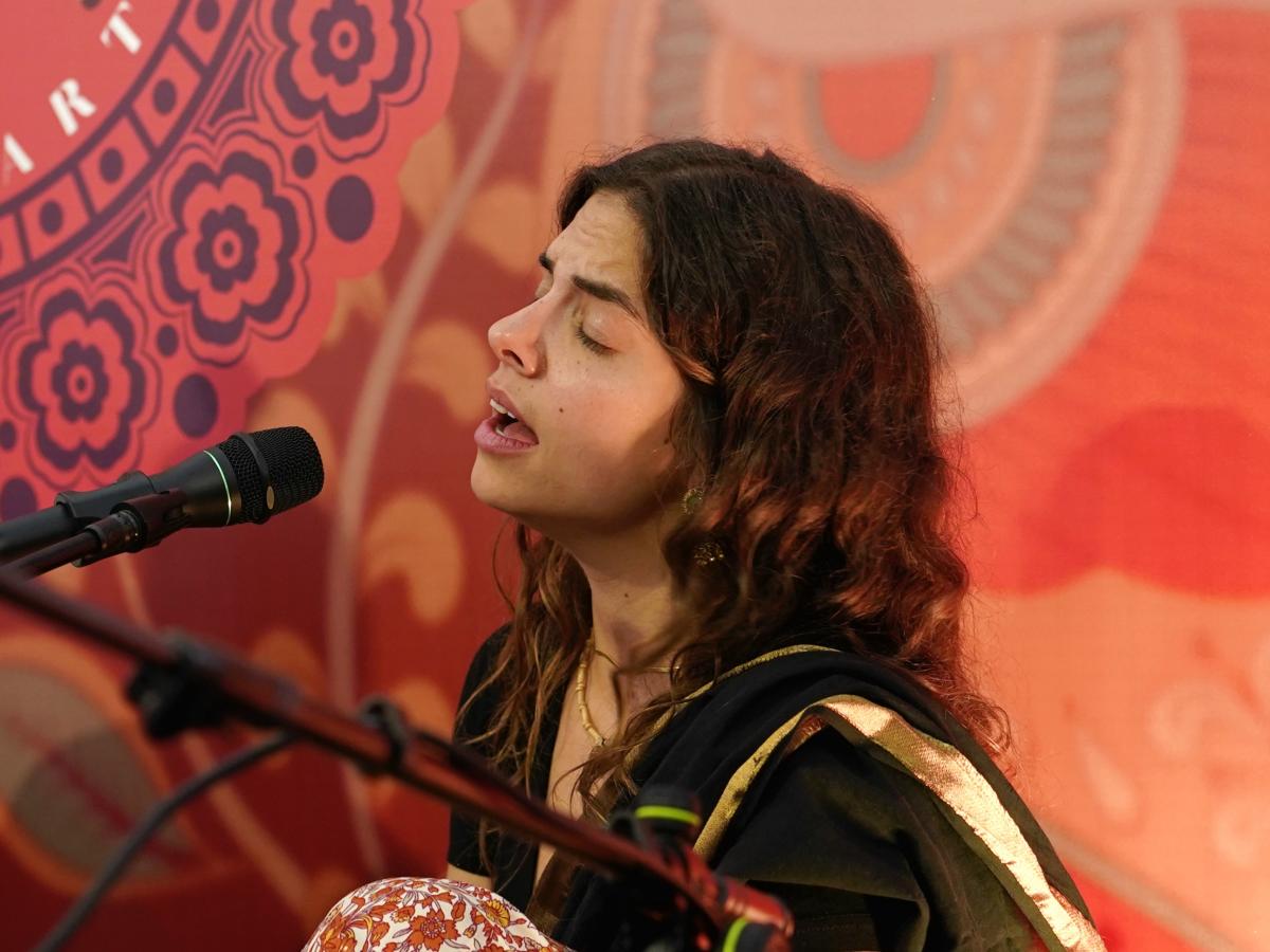 Stefanie Kümmerle hockt auf dem Boden. Eines ihrer Knie ist zu sehen und dass sie eine bunte Blumenhose trägt. Ihre Oberbekleidung ist schwarz. Sie hat braune offene lange lockige Haare. Ihre Augen sind geschlossen, ihre Stirn gerunzelt und sie singt hingebungsvoll in ein Mikrophone. Hinter ihr die Wand ist rot-orange mit Ranken und Blumen.