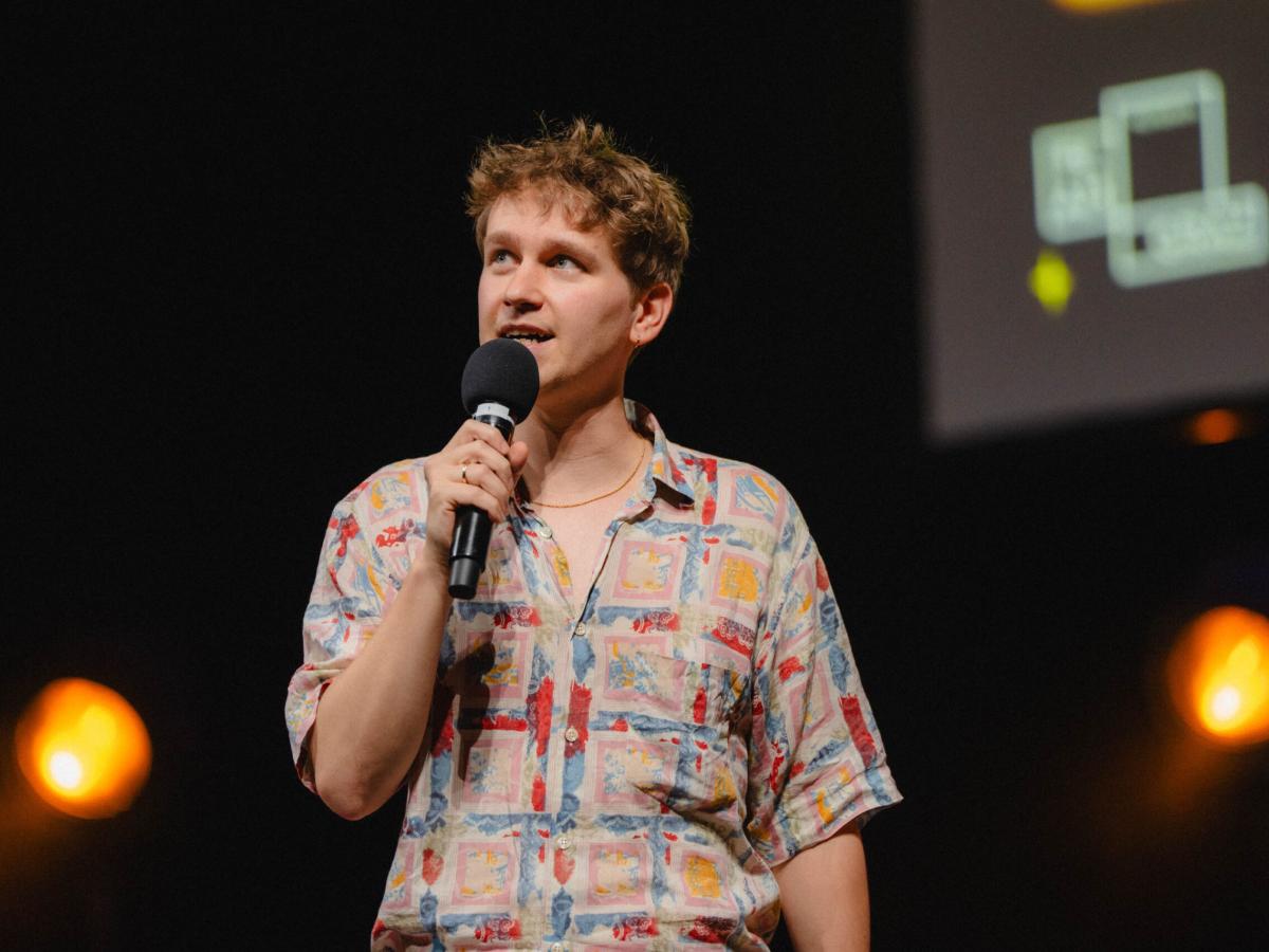 Yannick Steinkellner, dunkelblonde kurze gelockte Haare, trägt ein bunt gemustertes 90er Jahre Hemd mit kurzen Ärmeln. Das Hemd ist weit aufgeknöpft und es ist eine feine Kette auf seiner Haut zu sehen. Er schaut rechts nach oben und spricht in ein Mikrophone.