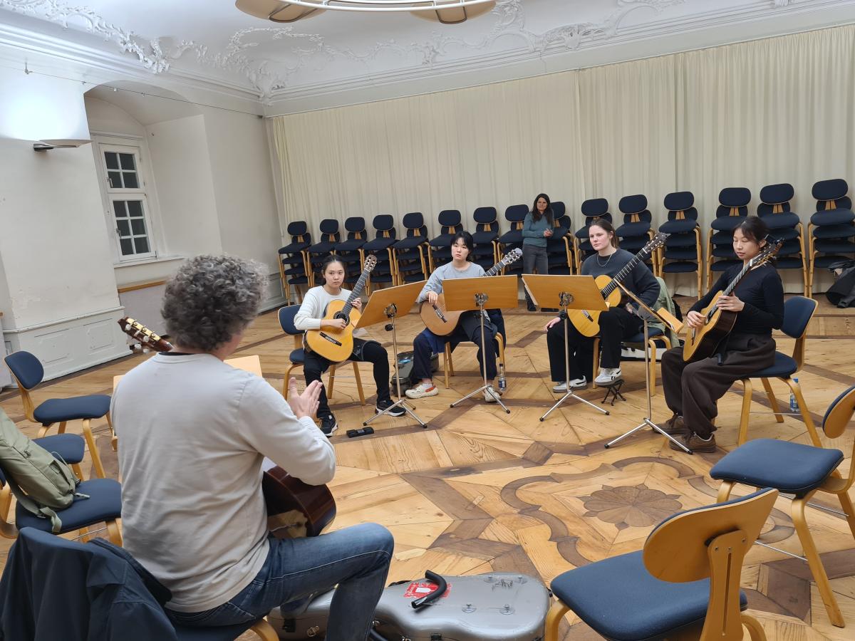 Vier Gitarristinnen beim Üben mit Prof. Tillmann Reinbeck im Stuhlkreis. Die Dozentin für Gesundheit / Prävention schaut im Hintergrund zu.