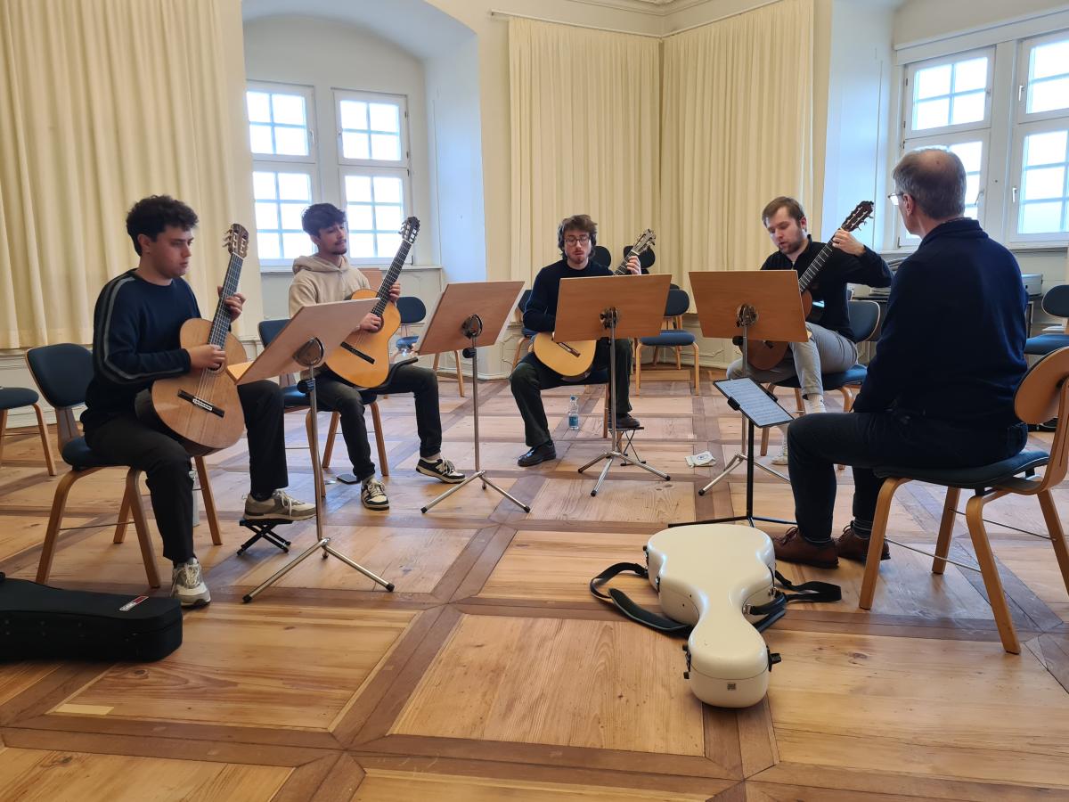 Vier Gitarristen beim Üben im Stuhlkreis. Der Dozent sitzt ihnen gegenüber. Man sieht ihn nur von hinten.