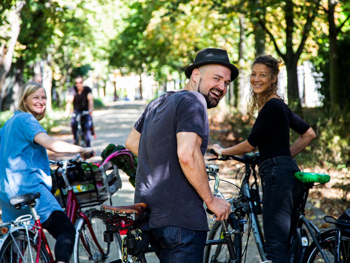 drei menschen auf Fahrrädern in einer grünen Allee. Sie schauen freundlich in die Kamera.