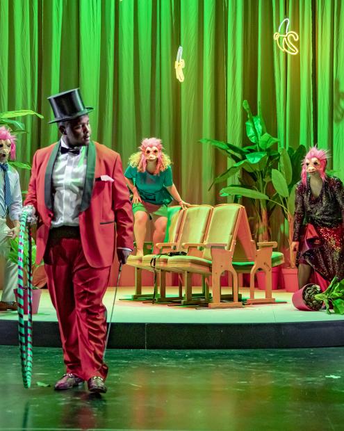 Don Giovanni: vier Studierende auf der Bühne. Im Vordergrund ein Student in Anzug , Gehstock und einem Hoola-Hoop-Reifen in der Hand. Im Hintergrund auf einem Rondell drei Studierende mit Masken auf den Gesichtern.