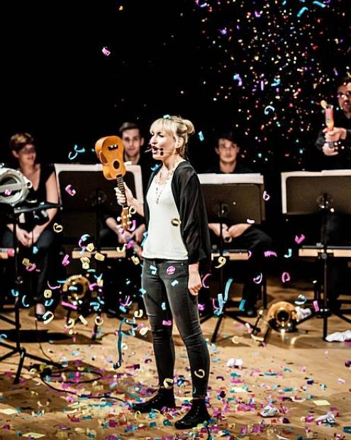 Frau singt mit Okulele falschherum in der Hand. Hinter ihr Menschen an Notenständern. Sie steht im Konfettiregen.