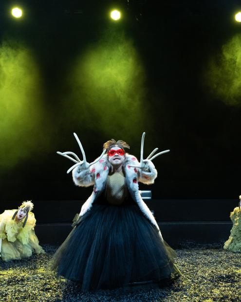 Mann mit sehr langen Fingern, Tüllkleid udn Pelzmantel steht in der Mitte. Um ihn herum sitzen gelb gekleidete ungläubig guckende Personen.