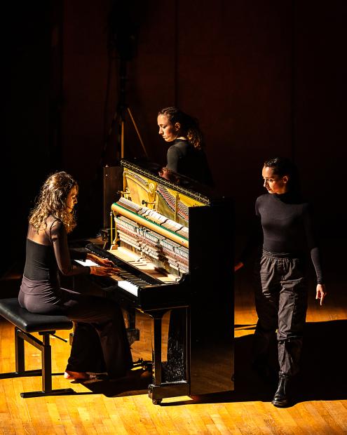 Eine Studentin spielt barfuß Klavier, zwei Studentinnen schauen zur Pianisten. Alle drei sind schwarz gekleidet.