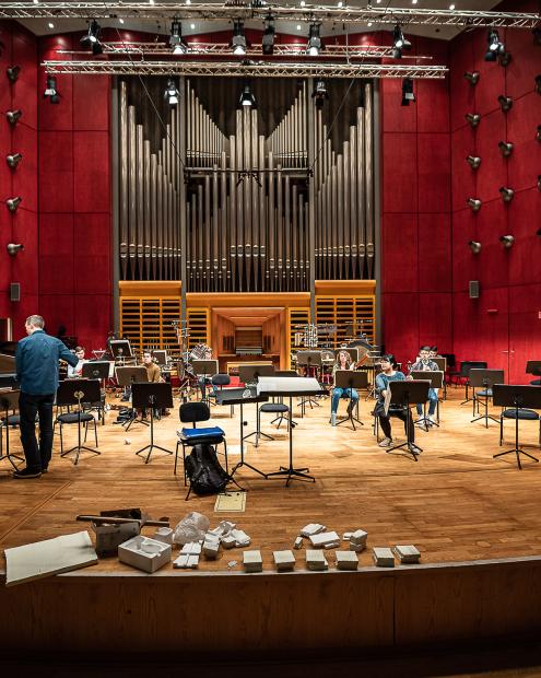 Blick vom Zuschauerraum auf die Bühne des Werkstattfestivals. Christ M Löser ist im Gespräch. An der Bühnenkante liegen viel kleinere, weiße Styroporstapel. Das Podium ist mit einem Orchesteraubfau für ein Kammerensemble versehen.