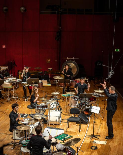 Studierende an unterschiedlichen Schlagwerken auf der Bühne, dirigiert von Christof M Löser.
