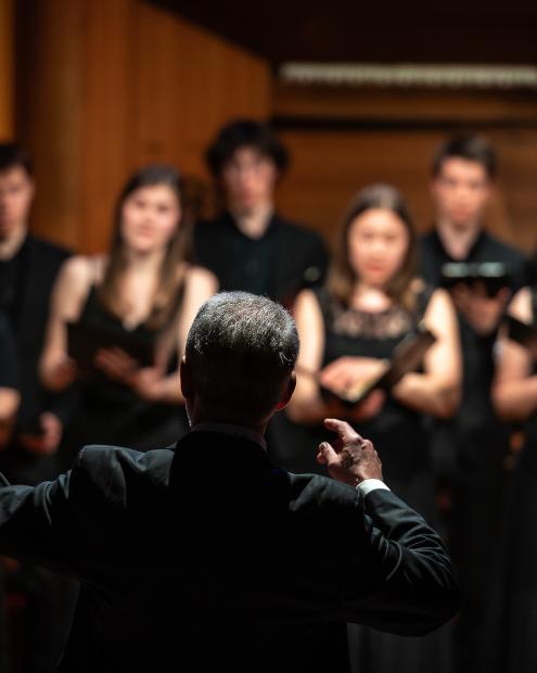 Prof. Denis Rouger dirigiert den Kammerchor. Der Fokus liegt auf seiner Person. Die Mitglieder sind nur teilweise zu erkennen.