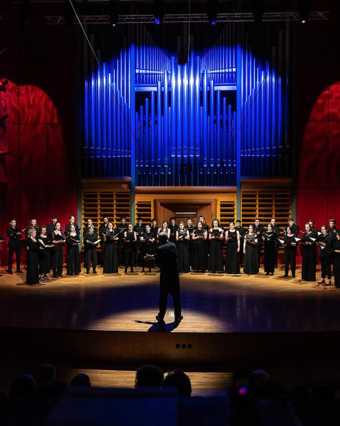 Der Kammerchor unter der Leitung von Prof. Denis Rouger im Konzertsaal. Die Seitenwände und das Orgelprospekt sind atmosphärisch beleuchtet.