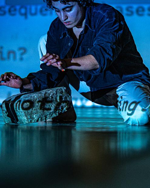 Knieende Studentin greift nach einem Felsbrocken, der vor ihr liegt. Die Umgebung ist in blaues Licht gefärbt, im Hintergrund sieht man Projektionen an der Wand.