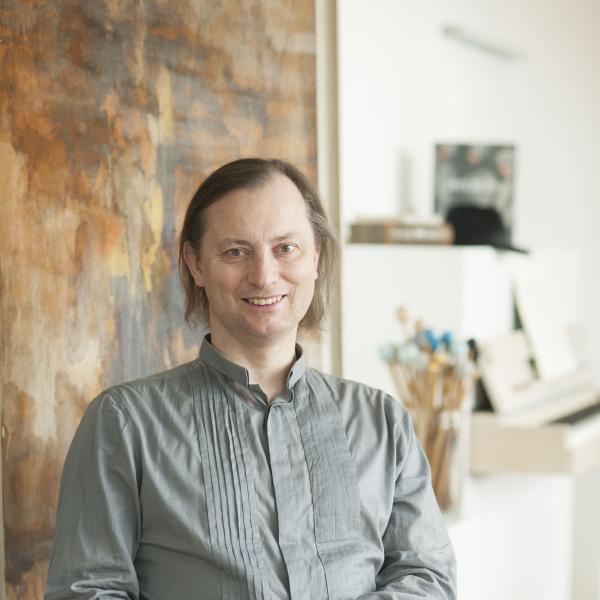 Klaus Dreher vor einem abstrakten, in Bastellfarben gehaltenen Bild auf einem grauen Sessel sitzend, im Hintergrund ein weißlackiertes Klavier.ier im Hintergrund