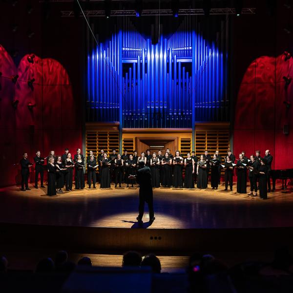 Der Kammerchor unter der Leitung von Prof. Denis Rouger im Konzertsaal. Die Seitenwände und das Orgelprospekt sind atmosphärisch beleuchtet.