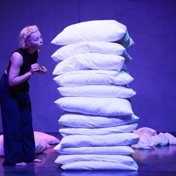 Szenenbild der Studierenden Eva Mario Hasler vor einem hohen weißen Kissenberg stehend und auf die Kissen pustend. Die Studierende ist in schwarz gekleidet und hat blonde Haare mit Undercut.
