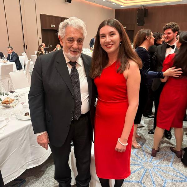 Aleksandra Domashchuk steht in einem roten Kleid und schwarzen Stiefeln neben einem älteren Mann im schwarzen Anzug und weißen Haaren. Beide lächeln in die Kamera.