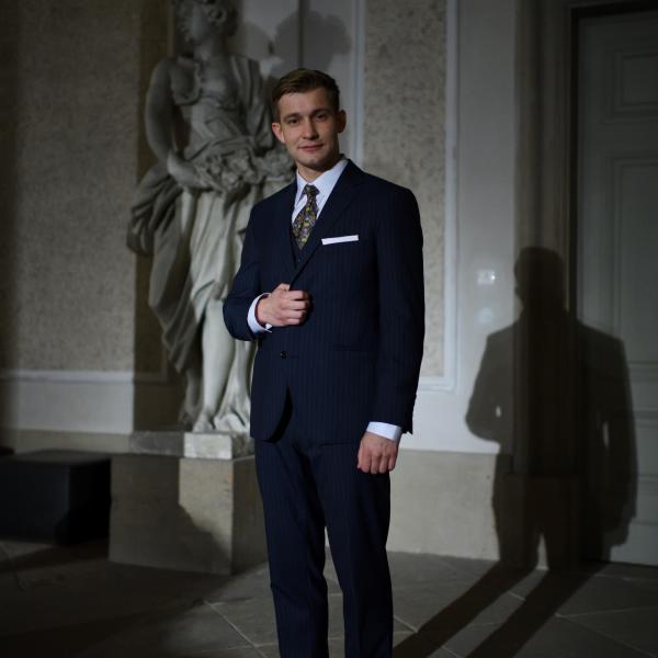 Daniel Domarecki steht in einem dunklen Anzug mit Kravatte und hellem Hemd vor einer Stein-Statue und lächelt in die Kamera.