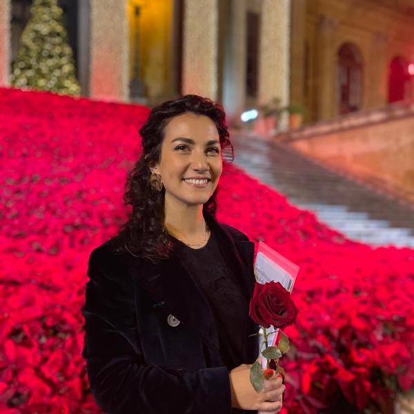 Alumna Eufemia Manfredi steht lachend mit einer roten Rose vor einem roten Rosen-Feld.