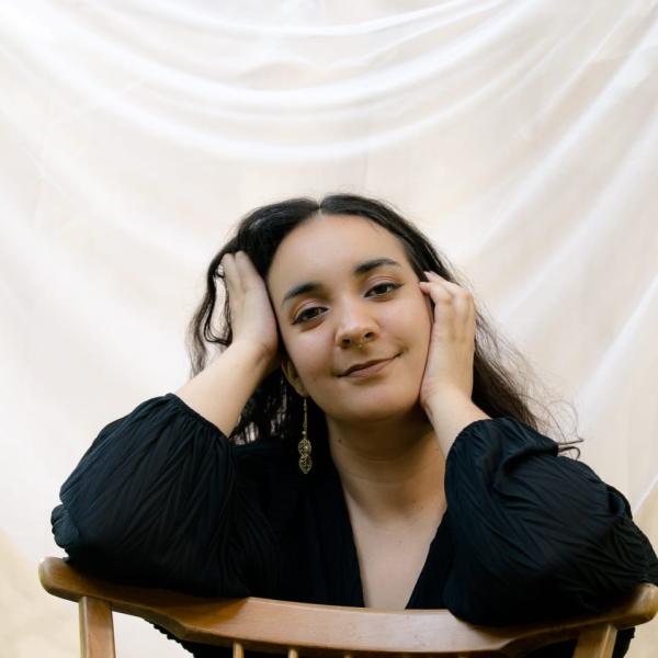 Inés López Fernández sitzt rittlinks auf einem Lehnstuhl und stützt ihren Kopf in beide Hände udn lächelt.