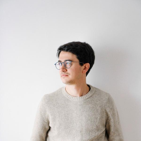 Giovanni Conti lehnt mit einem beigen Strickpulli und einer dunklen Hose an einer hellen Wand und schaut nach rechts in die Ferne. Er hat dunkle Haare, trägt eine Brille und hat seine Hände in seinen Hosentaschen.