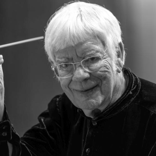 Portrait des Dirigenten Helmuth Rilling. Er hat kurze, weiße, dichte Haare und einen kurzen weißen Pony. Er trägt eine Brille, hat viele Falten im Gesicht, lächelt und hält in seiner rechten Hand einen Dirigentenstab.