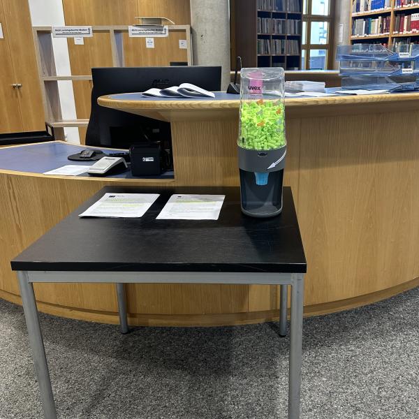 Das Bild zeigt eine Spenderbox, aus der man Ohrstöpsel entnehmen kann. Die Box steht auf einem Tisch an der Ausleihtheke der Bibliothek.