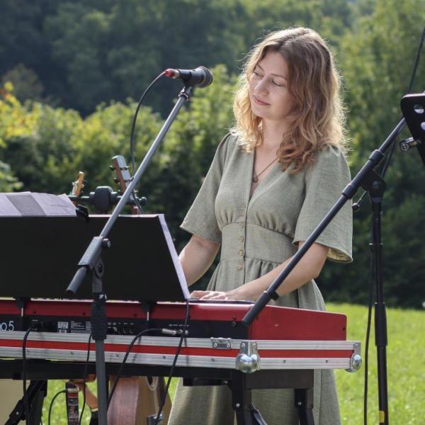 Studentin steht an einem Keyboard vor einem Mirkrophone in einem grünen Sommerkleid auf einer grünen Wiese udn hat die AUgen geschlossen.