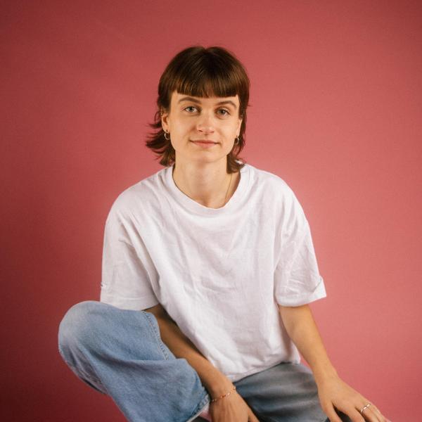 Foto der Sprechkunststudierenden Fabienne Dumont vor rotem Hintergrund in weißem T-Shirt und blauer lockerer Jeans.