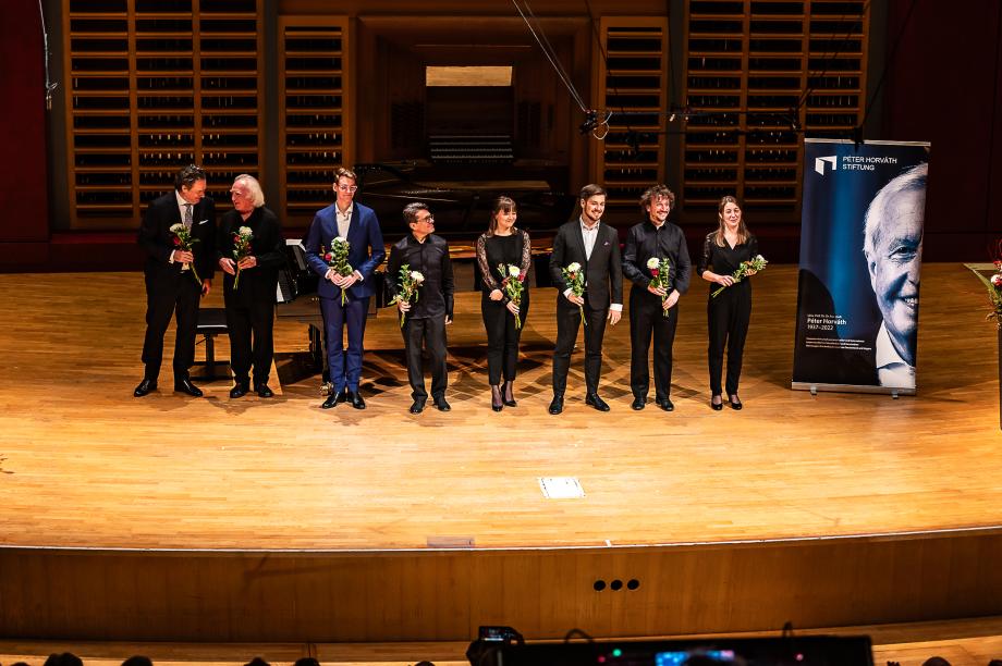 Mehrere festlich gekleidete Personen stehen während dem Abschiedsapplaus auf der Bühne, sie halten Blumen in der Hand.