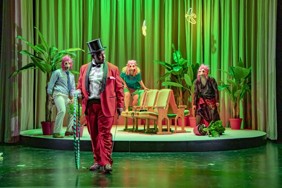 Don Giovanni: vier Studierende auf der Bühne. Im Vordergrund ein Student in Anzug , Gehstock und einem Hoola-Hoop-Reifen in der Hand. Im Hintergrund auf einem Rondell drei Studierende mit Masken auf den Gesichtern.