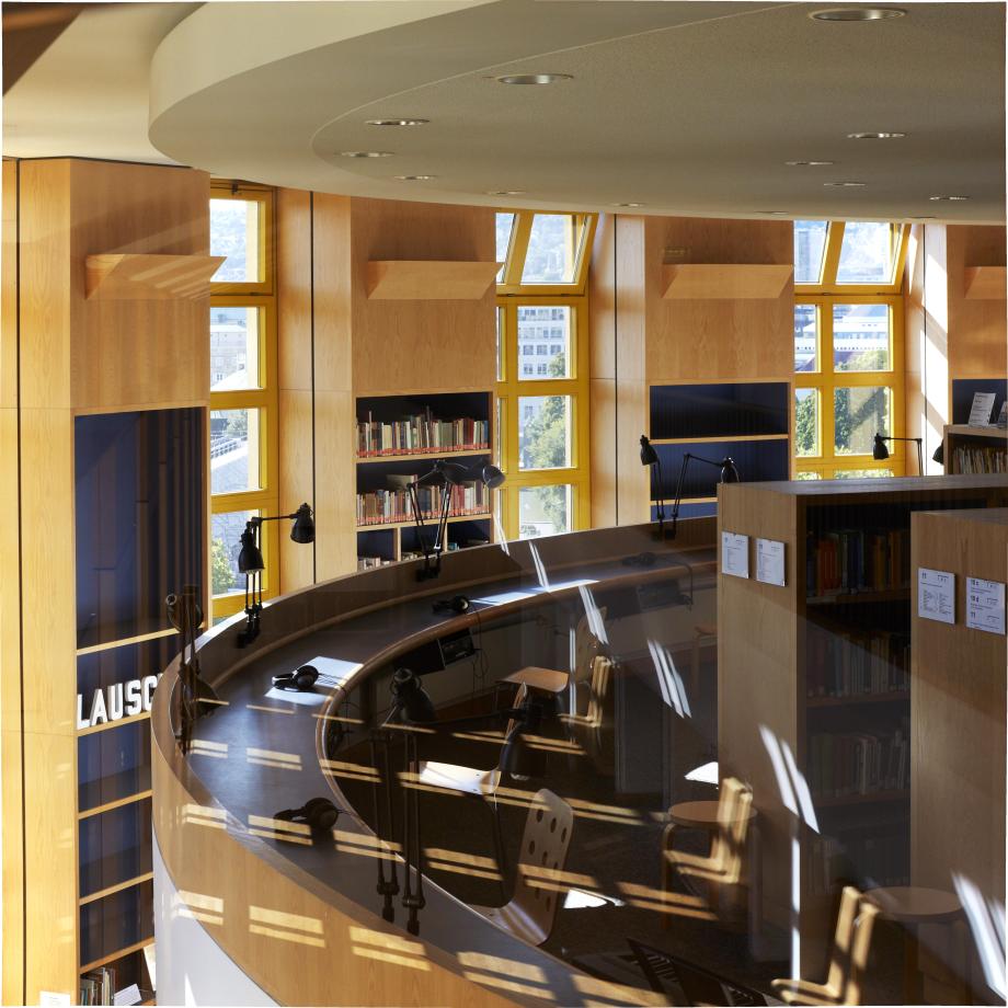 Auf dem Bild sind die Arbeitsplätze auf Ebene 9 zu sehen. Durch die Fenster fällt Licht auf die Tische, Stühle und Regale.