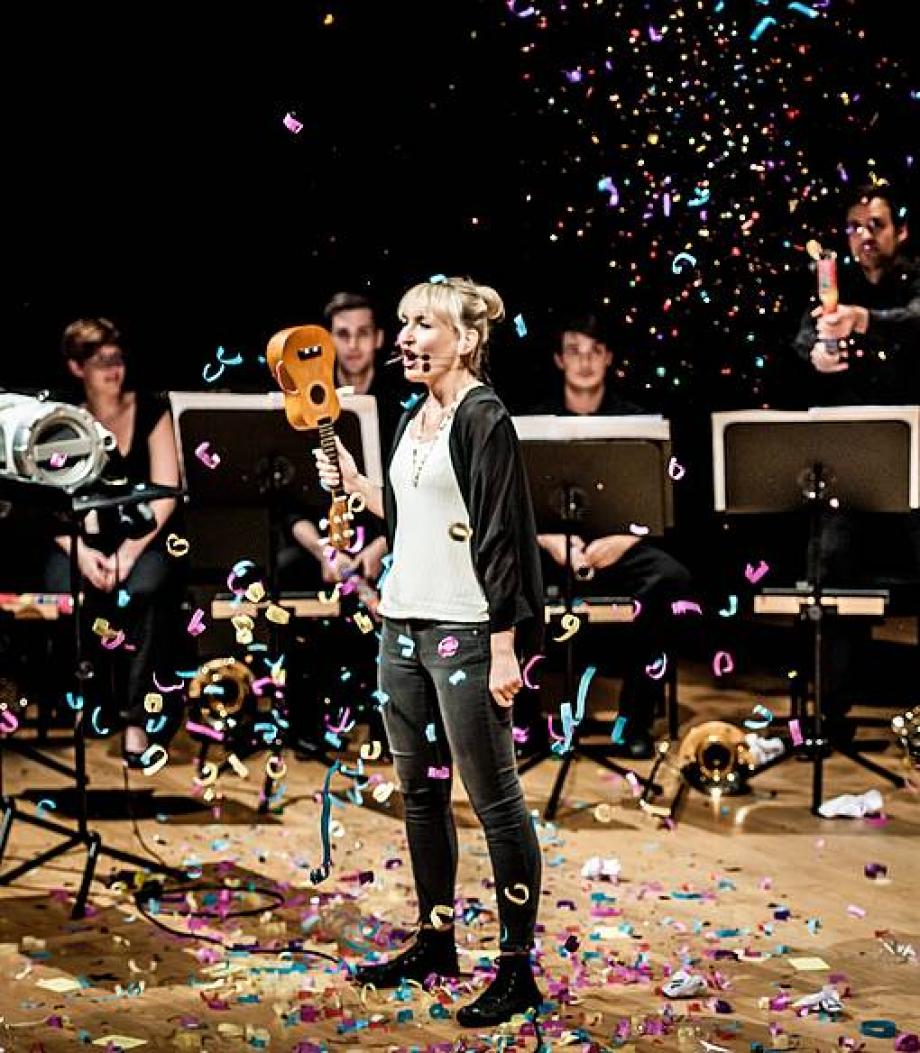 Frau singt mit Okulele falschherum in der Hand. Hinter ihr Menschen an Notenständern. Sie steht im Konfettiregen.