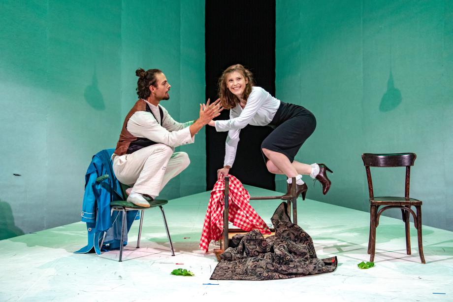 Mann hockt auf Stuhl, Frau auf umgedrehtem Tisch. Frau freudig, Mann schaut Frau beschwörend an.