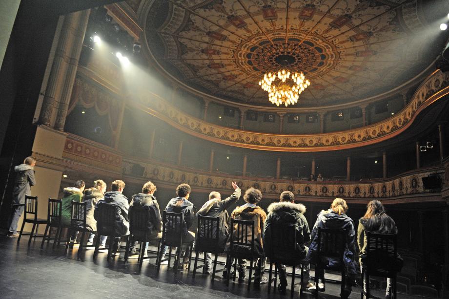 Meschen sitzen an Bühnenrand auf Stühlen mit Rücken zum Fotografen.