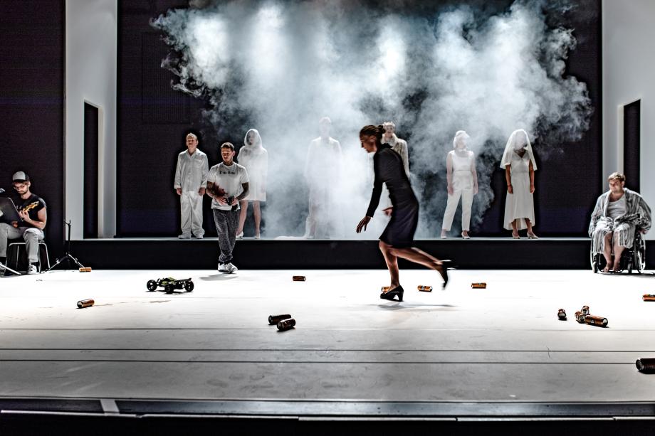 viele Menschen auf der Bühne, die im Nebel liegt. Auf dem Boden Dosen und ein Spielzeugauto.