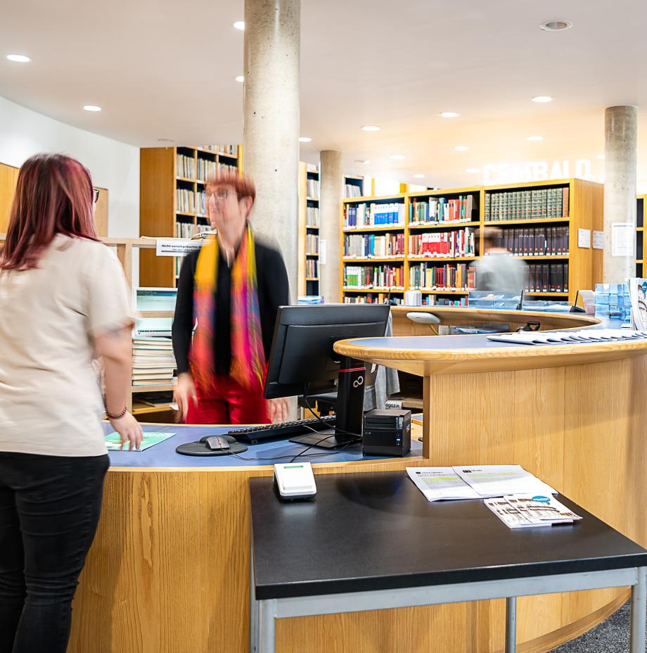 Das Bild zeigt die Theke in der Bibliothek. Zwei Personen stellen eine Ausleihsituation dar. Im Hintergrund sind Bibliotheksregale zu sehen.