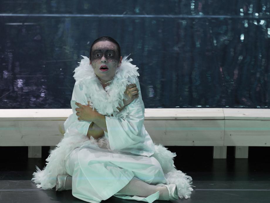 Studentin ganz in weiß gekleidet, mit einer Federboa umd en Hals, ihre Augenpartie ist schwarz geschminkt. Sie umarmt sich aufd em Boden sitzend selbst und schaut erschreckt mit offenem Mund.