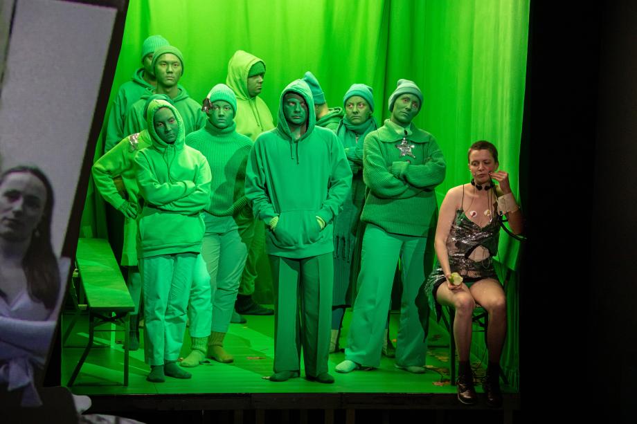 Bühnenszene mit Studierenden der Opernschule. Eine Frau mit kurzen Haaren sitzt im Vordergrund, hinter ihr steht eine Gruppe aus vollständig grünen Studierenden.