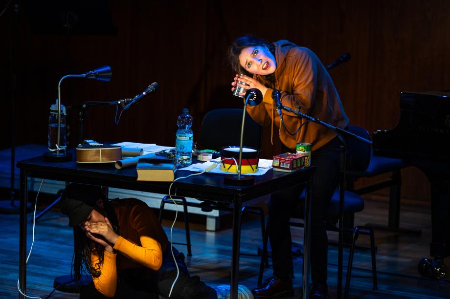Studierende hat eine Blechdose an ihrem Ohr und spielt "Stille Post". Um sie herum ein Tisch mit unterschiedlichen Requisiten, Mikrophonen und Lampen. Vor dem Tisch sitzt eine Studierende, die mit bei den Händen vor ihrem Gesicht ein Geräusch erzeugt.