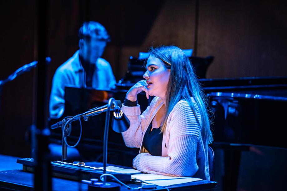 Studierende sitzt an einem Tisch und bildet mit der rechten Hand ein imaginäres Telefon nach. Im Hintergrund ist ein Pianist zu erkennen.