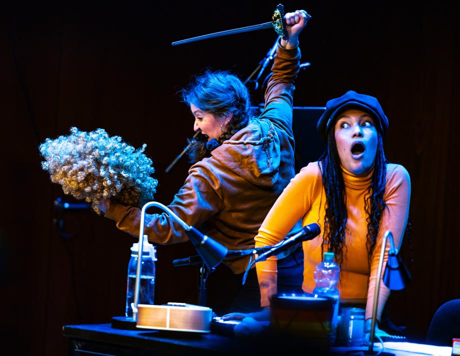 Eine Frau ersticht imaginär mit einem Dolch eine gelockte perücke. Die zweite Frau wendet sich mit Entsetzen von ihr ab.