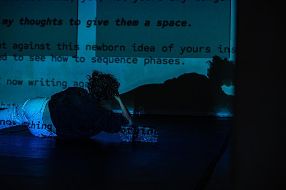 Frau liegt zu Betrachter mit dem Rücken auf der Seite und stützt ihren Kopf. Über die Leinwand vor ihr läuft ein Text.