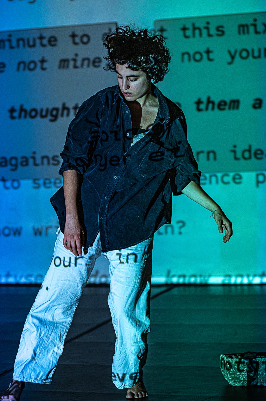 Blau illuminierte Studierende richtet ihren Blick auf den schwarzen Tanzboden. Im Hintergrund wird ein englischsprachiger Text an die Wand projiziert.