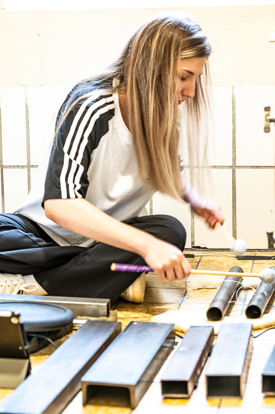 Im Schneidersitz spielende Schlagzeugerin. Verschiedene Becken, Stahlrohe und Stahlbleche umgeben sie.  