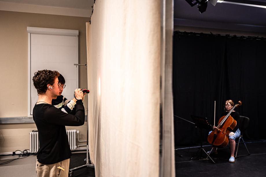 Zwei Studierende beleuchten eine Stoffwand mit Taschenlampen. Im Hintergrund sitzt eine Cellistin.