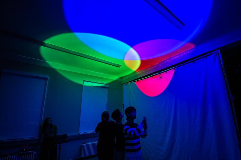 Ein in blau illuminierter Raum. Studierende beleuchten mit verschieden farbigen Taschenlampen Wand und Decke.