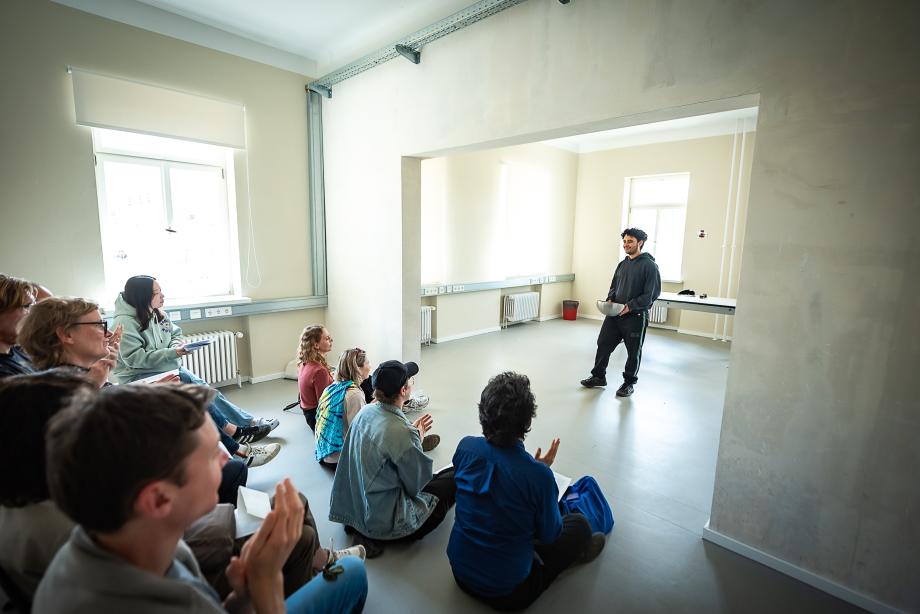 Studierender mit großer Alumniumschale interagiert mit dem sitzenden und stehenen Publikum. Der Raum ist von vielen Fenstern lichtdurchflutet.