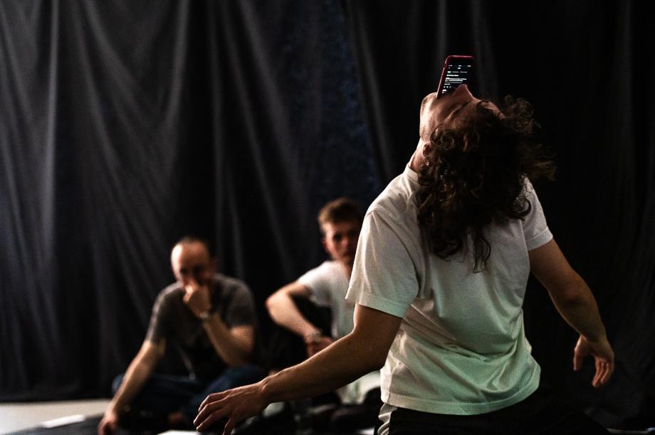 Blick von der Bühnen auf das Publikum. Ein Studierender hat die Hände seitlich ausgebreitet und ein rotes Smartphone im Mund.