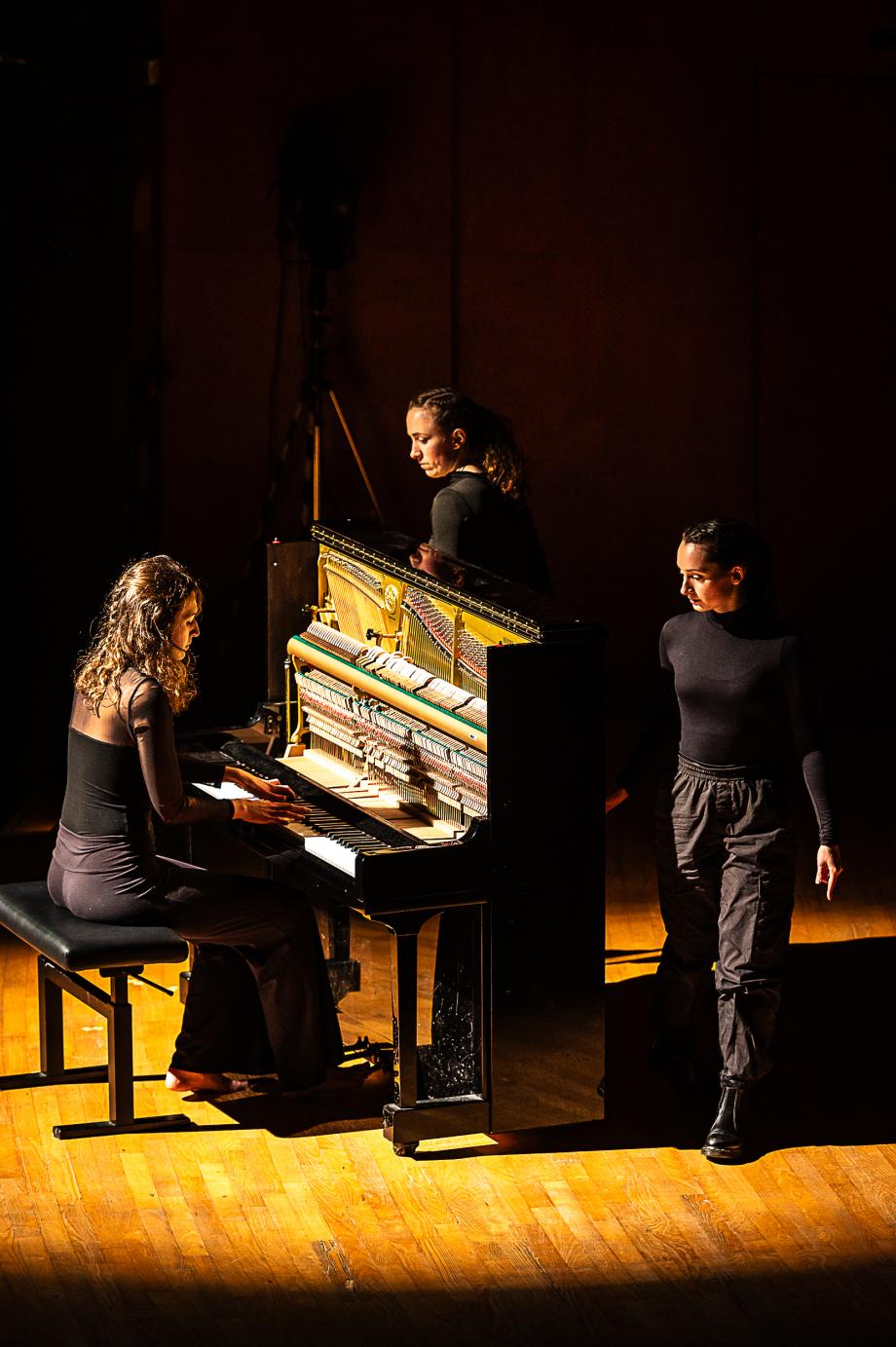 Eine Studentin spielt barfuß Klavier, zwei Studentinnen schauen zur Pianisten. Alle drei sind schwarz gekleidet.